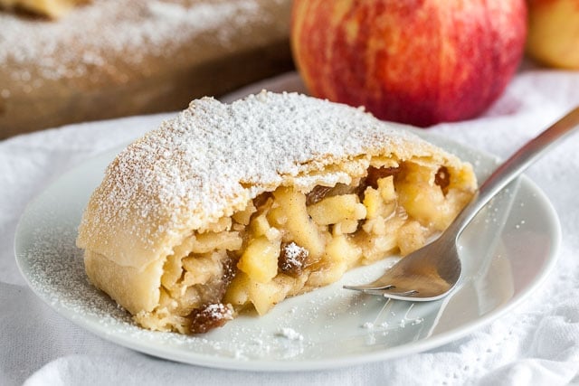 A piece of apple strudel topped with powdered sugar on a white plate with a fork on a white dishcloth, with an apple in the background.