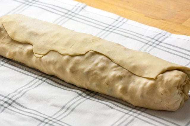 A bamboo cooking board with a white and grey plaid dishtowel and a rolled, unbaked apple strudel on top.