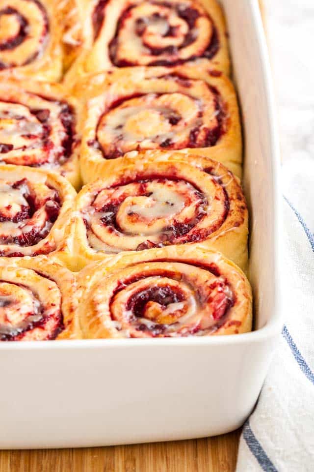 A white baking dish with orange cranberry rolls on a bamboo cutting board with a white and blue dishtowel.