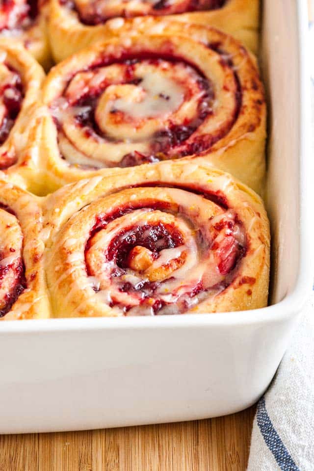 Close-up of a white baking dish with orange cranberry rolls on a bamboo cutting board with a white and blue dishtowel.