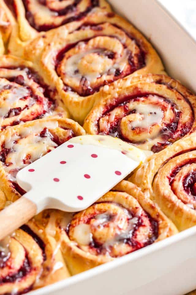 A white baking dish with orange cranberry rolls with sweet orange glaze with a white and red polka dot spatula on them.