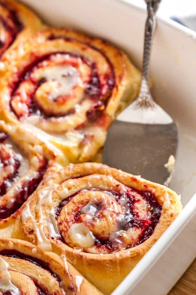 Close-up of a white baking dish with orange cranberry rolls. One of the rolls is missing and a cake server is lifting out another.