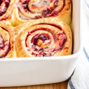 A white baking dish with orange cranberry rolls on a bamboo cutting board with a white and blue dishtowel.