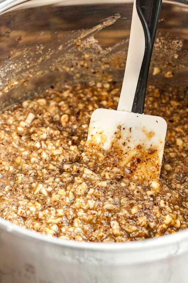 A stainless steel mixing bowl with caramelized nut topping with a white and black spatula in it.