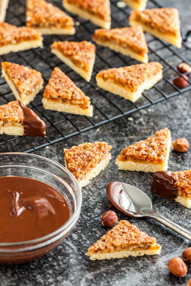 Nut corners on and next to a black cooling rack on a dark surface. Their corners have not yet been dipped into the glass bowl of molten chocolate.