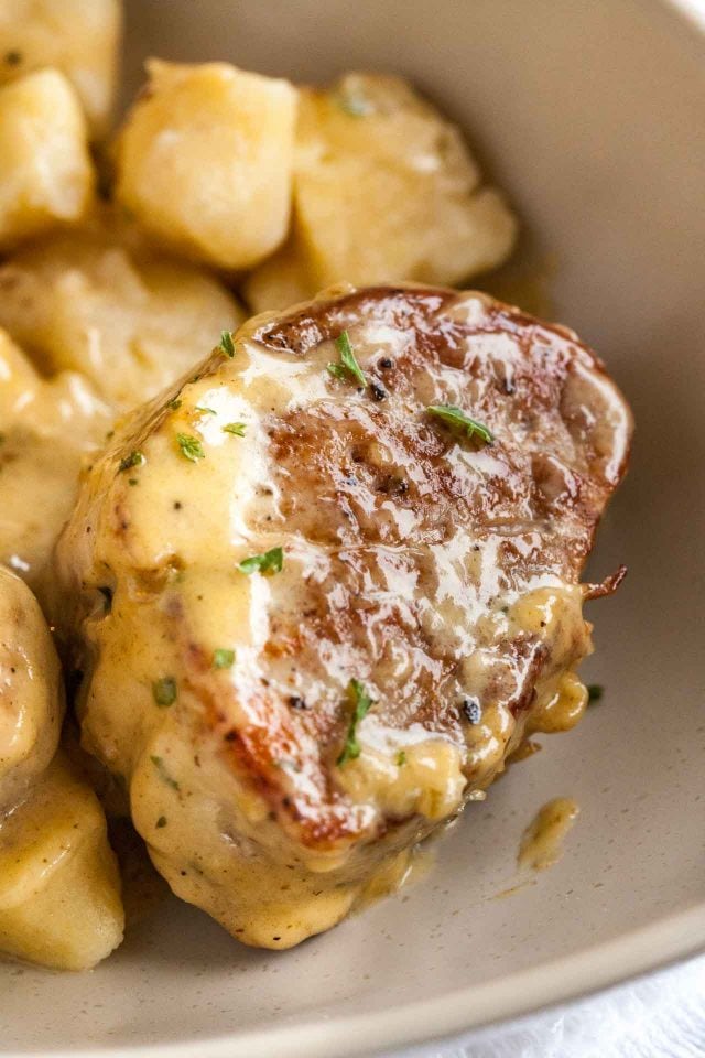 Close-up of a pan-seared pork medallion, covered in blue cheese sauce in a grey plate next to some potatoes.