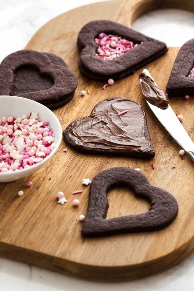 A wooden cutting board with heart shaped chocolate cookies in various stages of completion. In the foreground there are the top and bottom part of the cookie and a knife has applied nutella to the bottom. The top has a smaller heart cut out. There\'s a small bowl of sprinkles next to it.