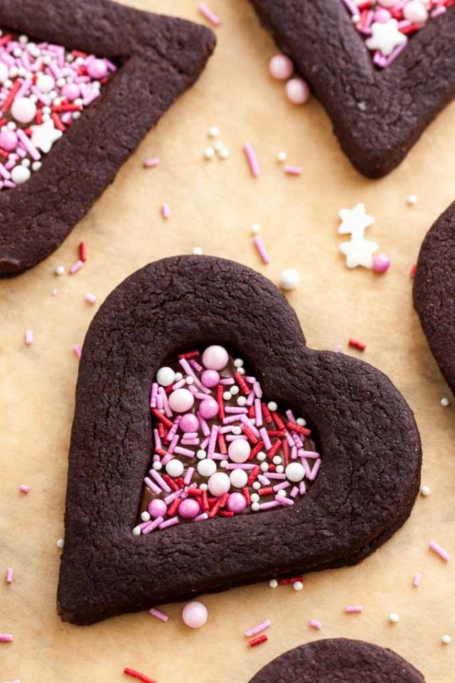 Top-down shot of heart-shaped chocolate cookies with a heart-shaped indentation filled with pink sprinkles on parchment paper with some sprinkles.