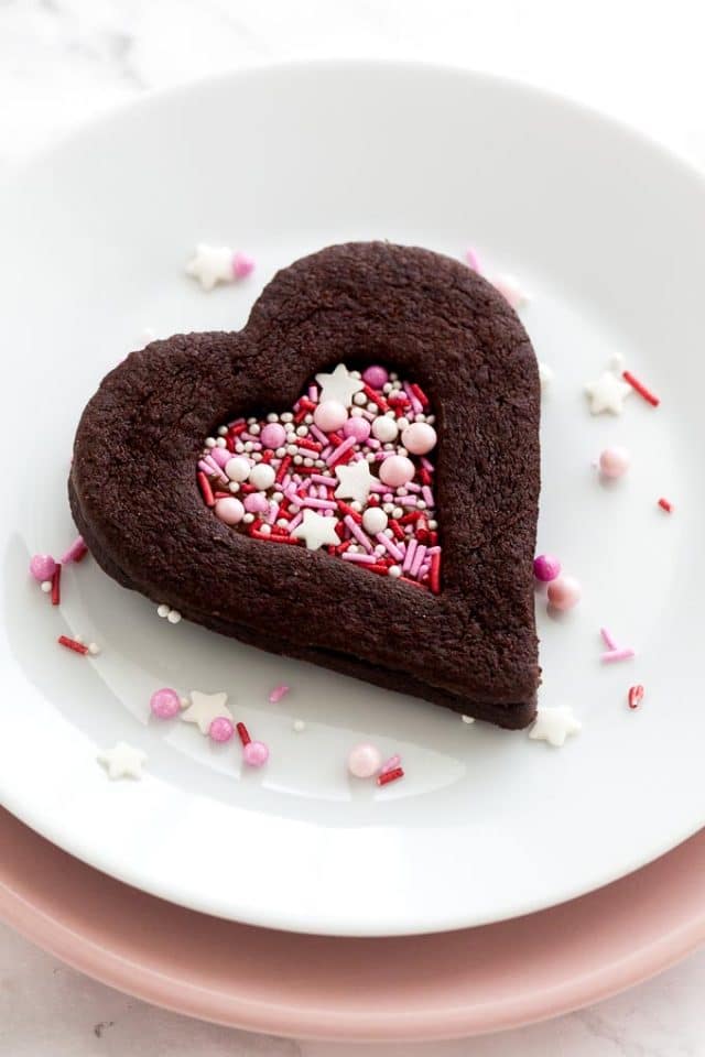 A heart-shaped chocolate cookie with a heart-shaped indentation filled with pink sprinkles on a white plate