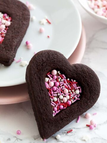 Close-up of a heart-shaped chocolate cookie with a heart-shaped indentation filled with pink sprinkles leaning against a stack of plates garnished with sprinkles. There's another cookie on the stack of plates and a small white bowl of pink sprinkles.