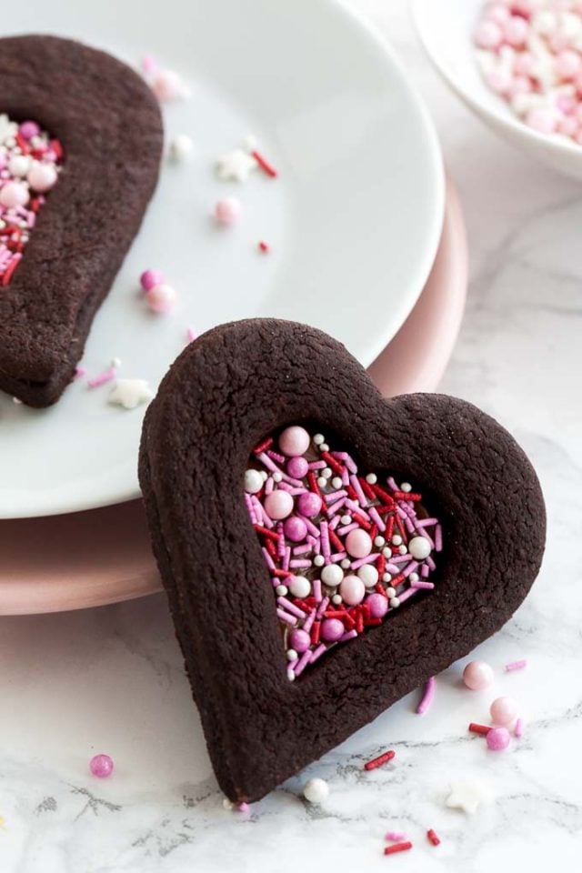 Close-up of a heart-shaped chocolate cookie with a heart-shaped indentation filled with pink sprinkles leaning against a stack of plates garnished with sprinkles. There\'s another cookie on the stack of plates and a small white bowl of pink sprinkles.