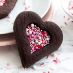 A heart-shaped chocolate cookie with a heart-shaped indentation filled with pink sprinkles leaning against a stack of plates garnished with sprinkles. There's another cookie on the stack of plates and a small white bowl of pink sprinkles.