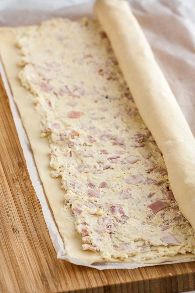 A layer of uncooked puff pastry with ham and cheese mixture on parchment paper lying on a wooden cutting board. The puff pastry is being rolled up from one side to form the pinwheels.