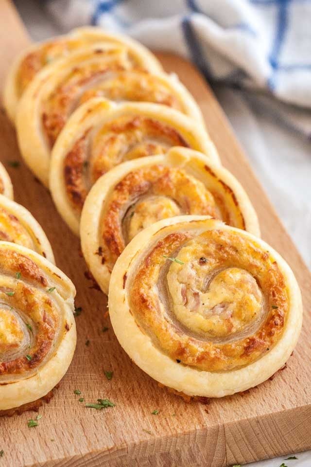 Two rows of ham and cheese pinwheels on a wooden cutting board next to a white and blue dishtowel.