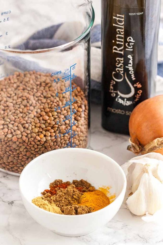 A glass measuring bowl with lentils, a squash, a bulb of garlic and a white bowl with spices on a marble surface with a white and blue dishtowel in the background.