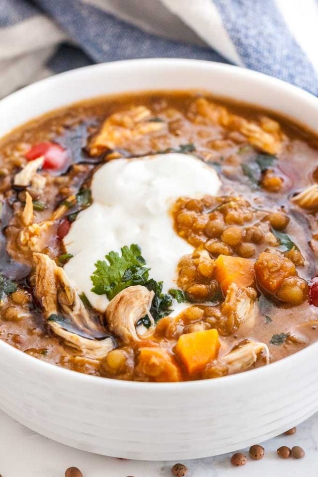 A white bowl of Lentil soup with vegetables and chicken garnished with parsley and sour cream. There\'s a white and blue dish towel in the background.