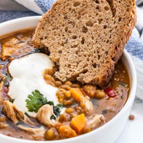A white bowl of Lentil soup with vegetables and chicken garnished with parsley and sour cream with a slice of bread. There's a white and blue dish towel in the background.