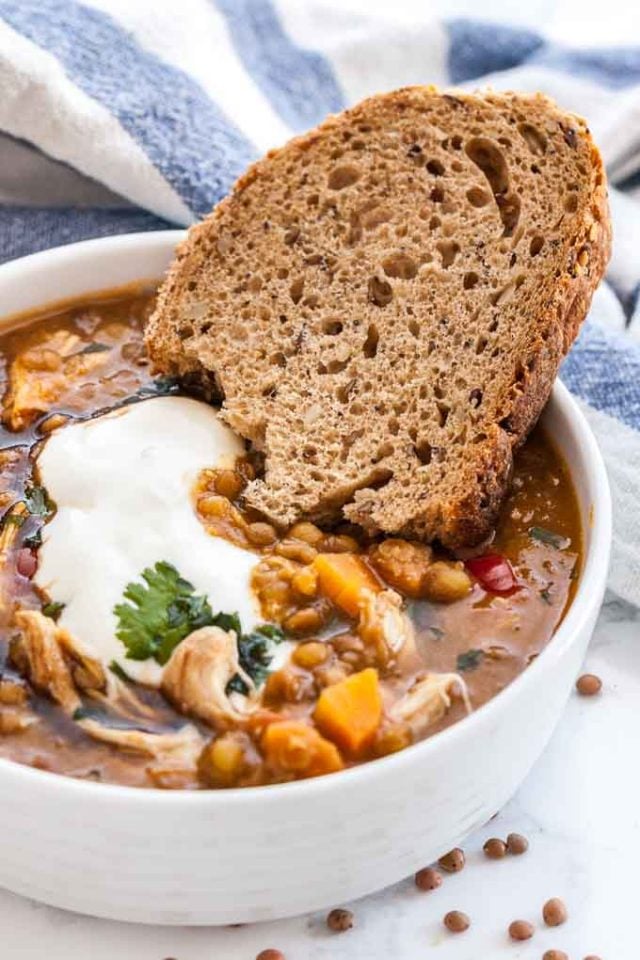 A white bowl of Lentil soup with vegetables and chicken garnished with parsley and sour cream with a slice of bread. There\'s a white and blue dish towel in the background.