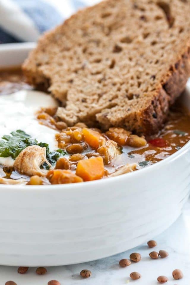 Close-up of a white bowl of Lentil soup with vegetables and chicken garnished with parsley and sour cream with a slice of bread.