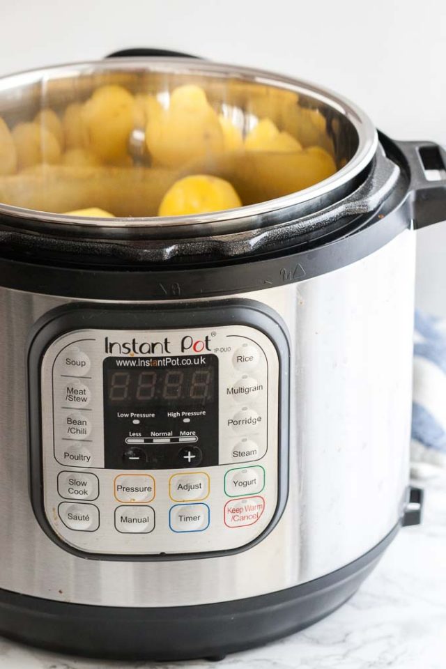 An instant pot with peeled potatoes next to a white and blue dish towel.