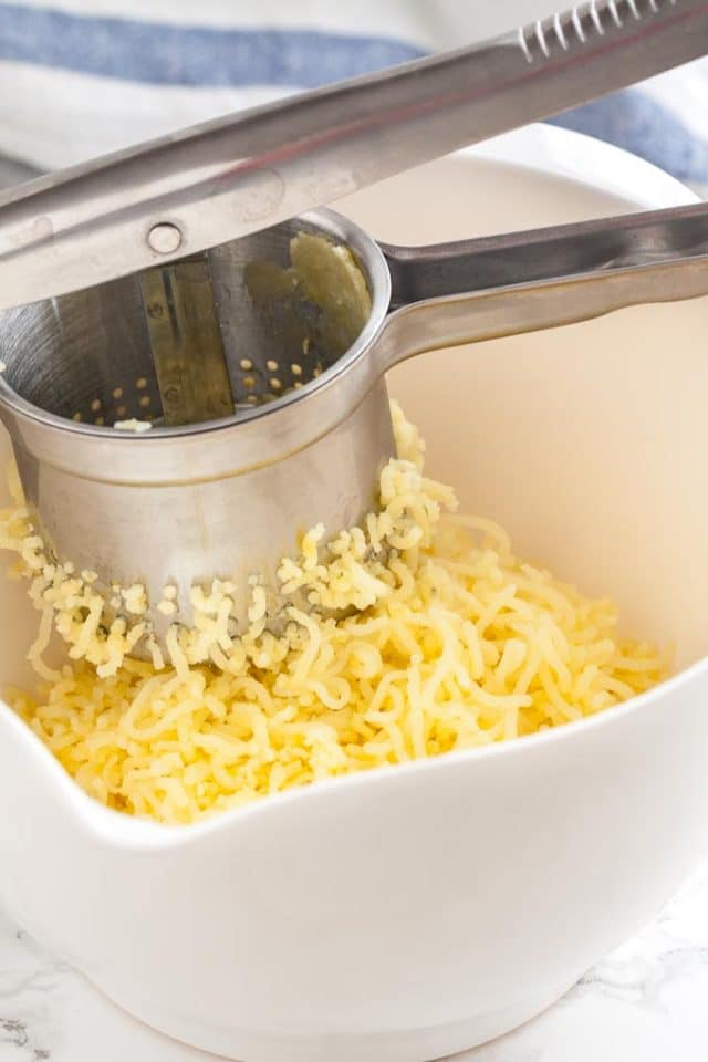 A white bowl with mashed potatoes. A potato ricer is squeezing potatoes into it.