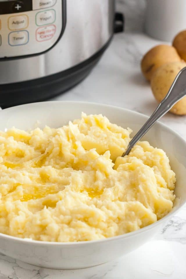 A white bowl of mashed potatoes with a spoon in it on a marble surface. There are potatoes and an instant pot in the background.