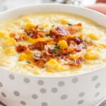 Close-up of a white bowl with silver dots of corn chowder topped with bacon bits and thyme.