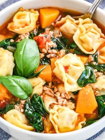 Close-up of a white bowl of spinach tortellini soup garnished with basil leaves.