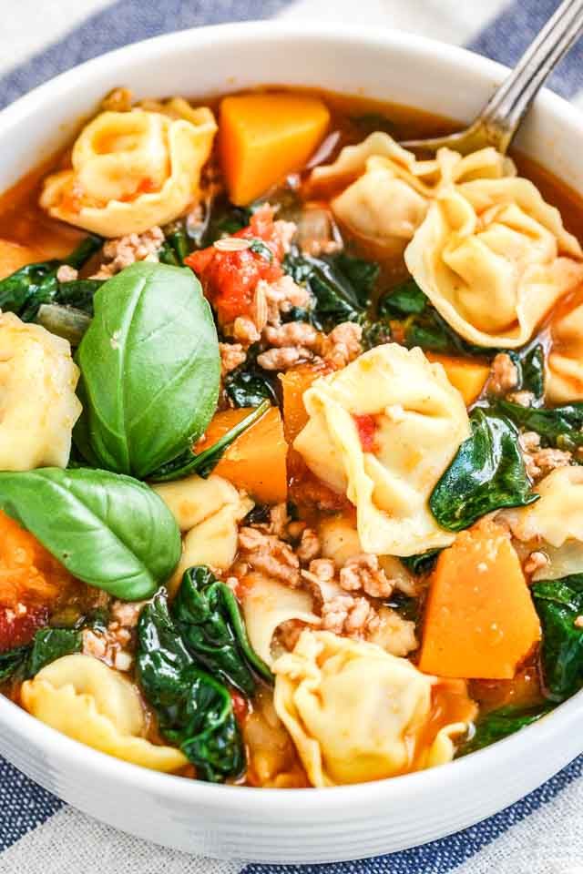 Close-up of a white bowl of spinach tortellini soup garnished with basil leaves.