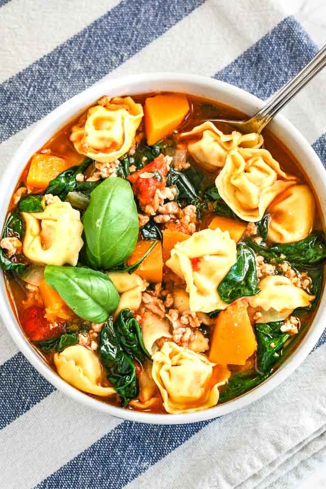 Top-down shot of a white bowl of spinach tortellini soup garnished with basil leaves on a white and blue dishtowel.