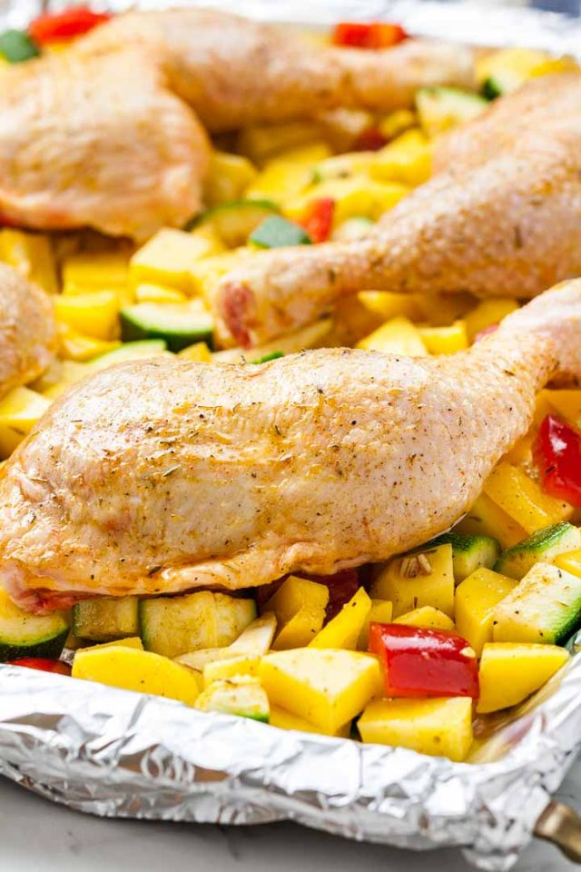 A baking sheet lined with aluminum foil, with vegetables (peppers, potatoes and zucchini) and chicken legs on top.