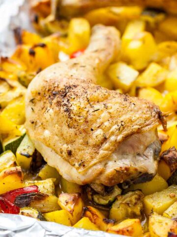 Close-up of a baked chicken leg on some roasted vegetables(peppers, potatoes and zucchini) on a grey plate next to a white and blue dishtowel.