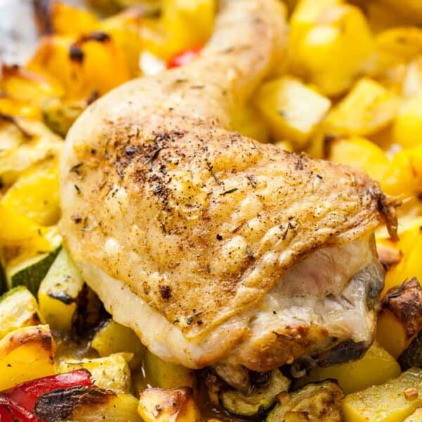 Close-up of a baked chicken leg on some roasted vegetables(peppers, potatoes and zucchini) on a grey plate next to a white and blue dishtowel.