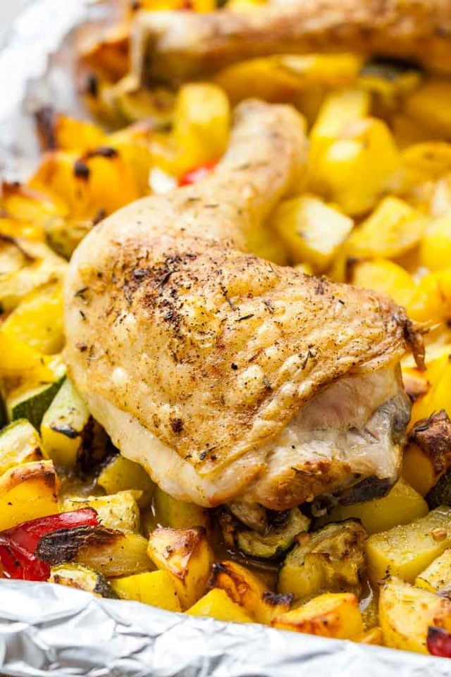 Close-up of a baked chicken leg on some roasted vegetables(peppers, potatoes and zucchini) on a grey plate next to a white and blue dishtowel.