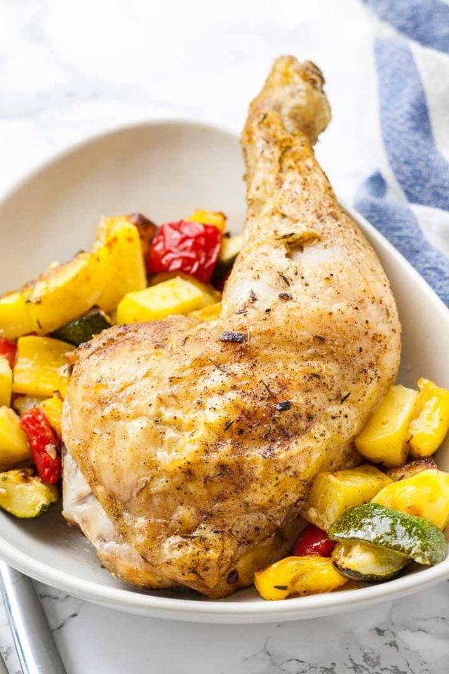 A baked chicken leg on some roasted vegetables (peppers, potatoes and zucchini) on a grey plate next to a white and blue dishtowel.