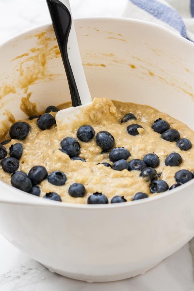 A white bowl of batter with blueberries and a black and white spatula in it.