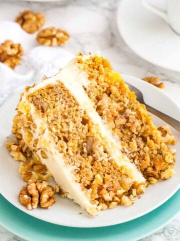 A slice of carrot pineapple cake garnished with walnuts lying on its side on a white plate stacked on a teal plate with a fork.