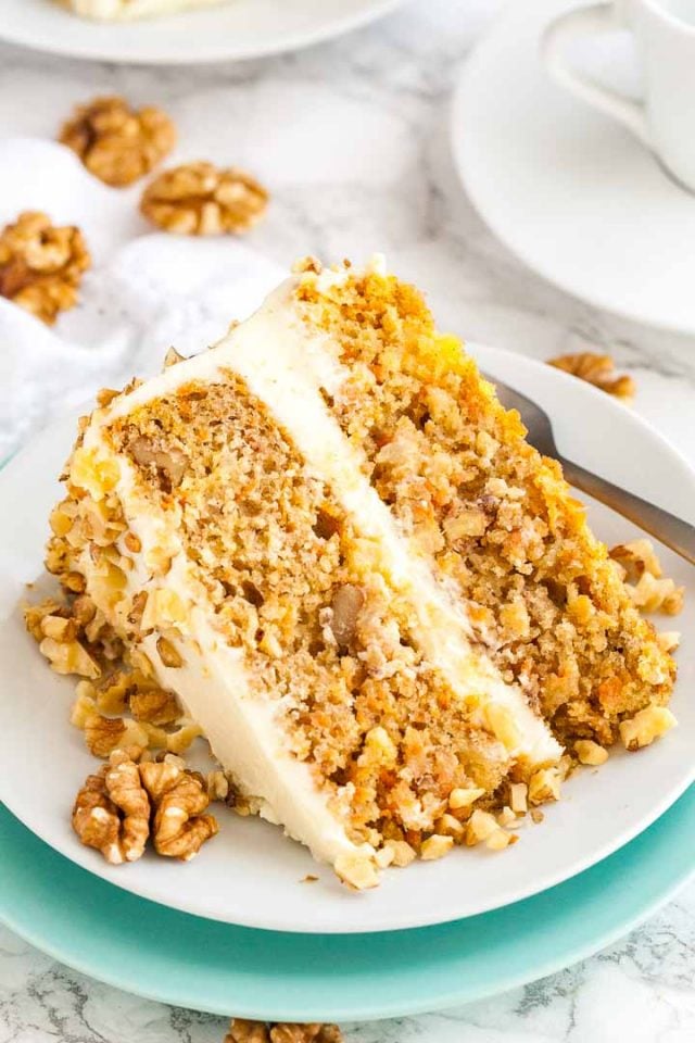 A slice of carrot pineapple cake garnished with walnuts lying on its side on a white plate stacked on a teal plate with a fork. 