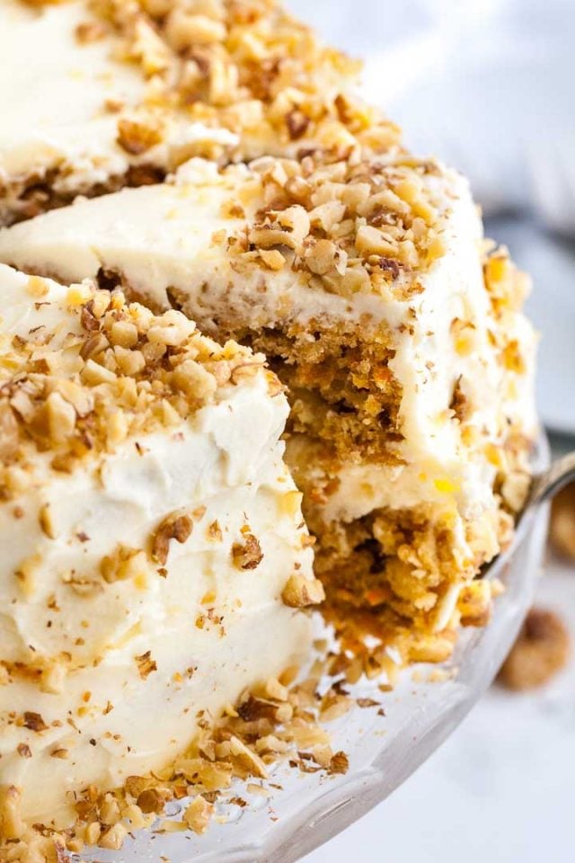 A carrot pineapple cake on a glass serving platter. A cake server is lifting out a slice.