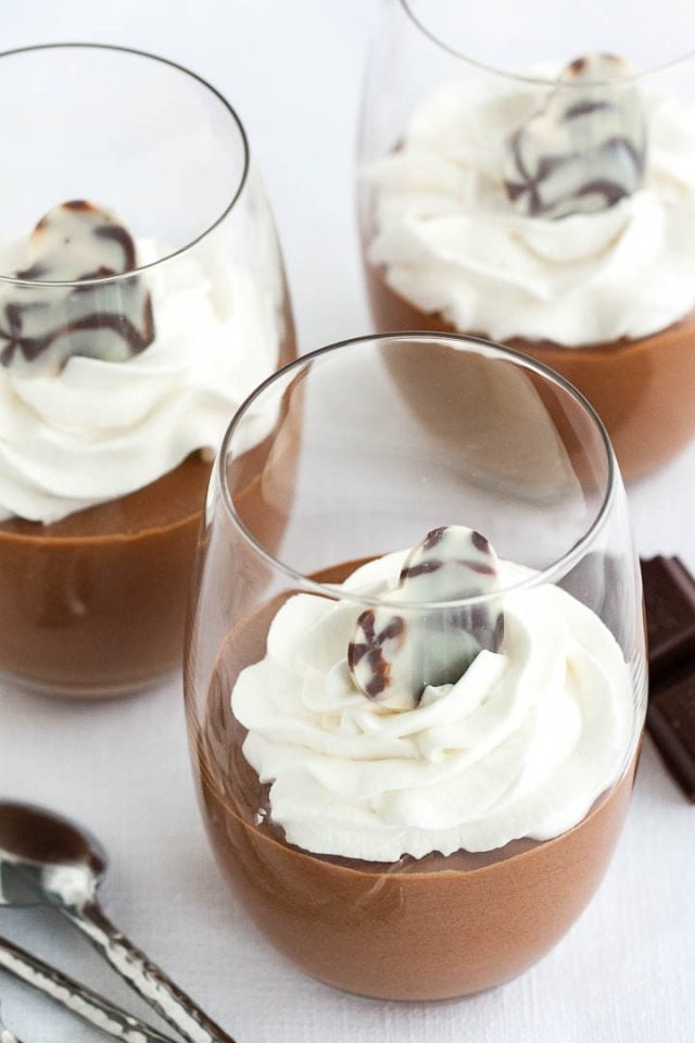Three glasses of chocolate mousse, topped with whipped cream next to pieces of chocolate and a spoon.