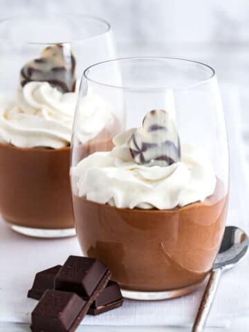 Chocolate mousse in a glass, topped with whipped cream next to pieces of chocolate and a spoon.