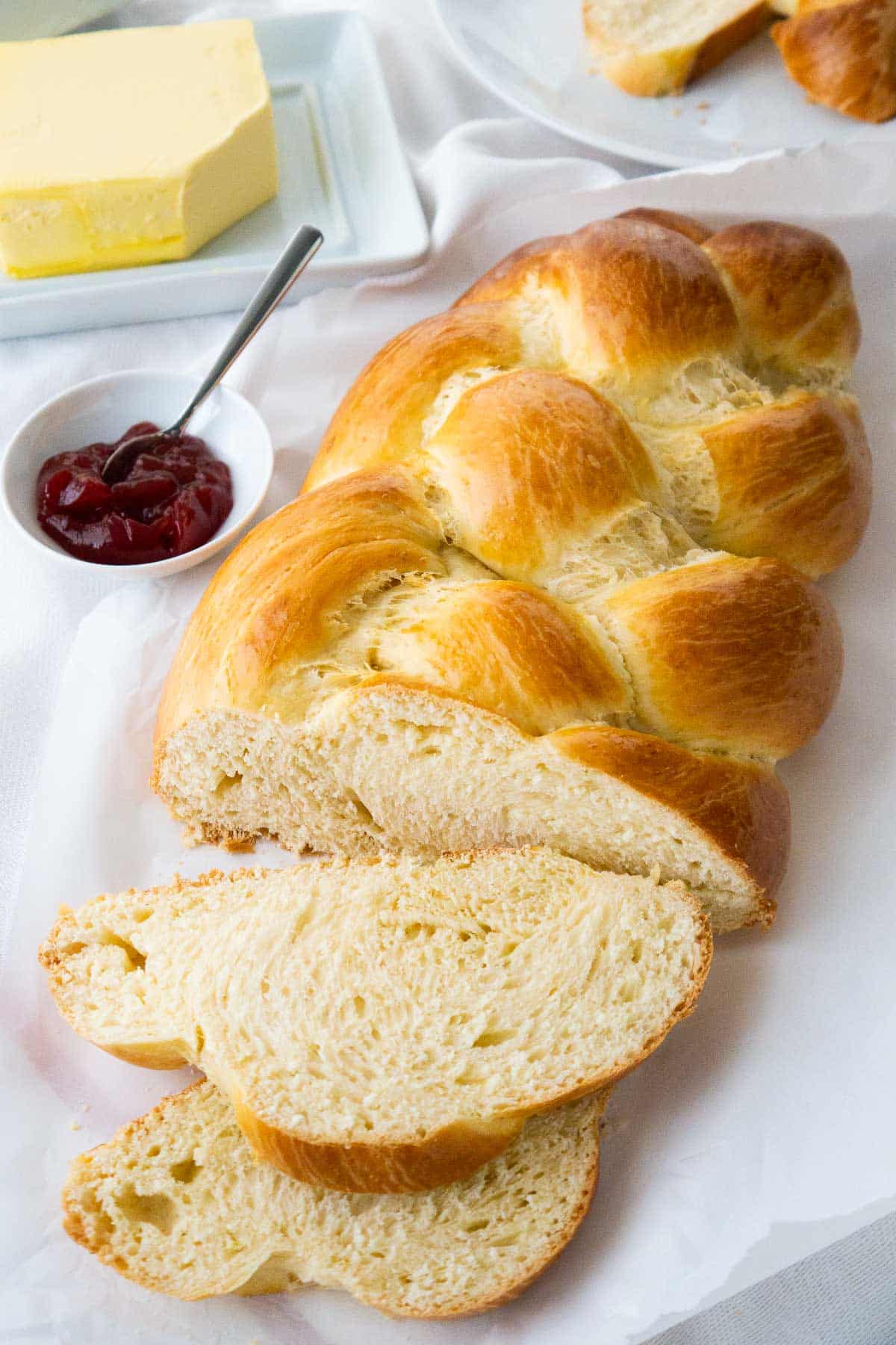 A loaf of German Sweet Bread, with a slice cut off and lying in front of it on a white dish towel next to a small white bowl of strawberry jam with a spoon in it.