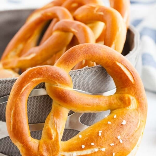 A pretzel leaning against a bread basket, containing more pretzels with a white and blue dish towel in the background.