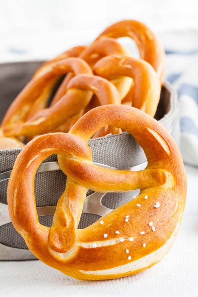 A pretzel leaning against a bread basket, containing more pretzels with a white and blue dish towel in the background.