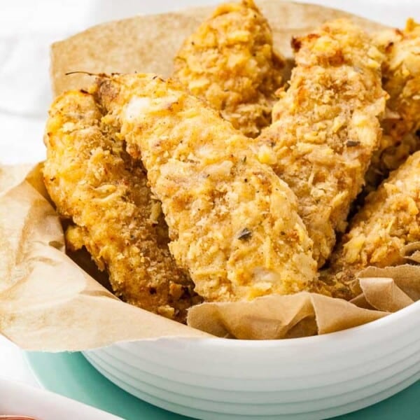 A white bowl lined with parchment paper, containing fried chicken tenders stacked on a teal plate.