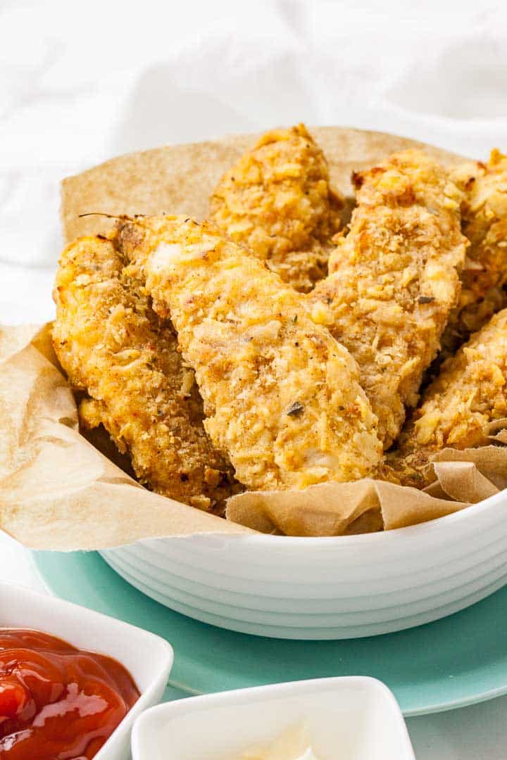 A white bowl lined with parchment paper, containing fried chicken tenders stacked on a teal plate.