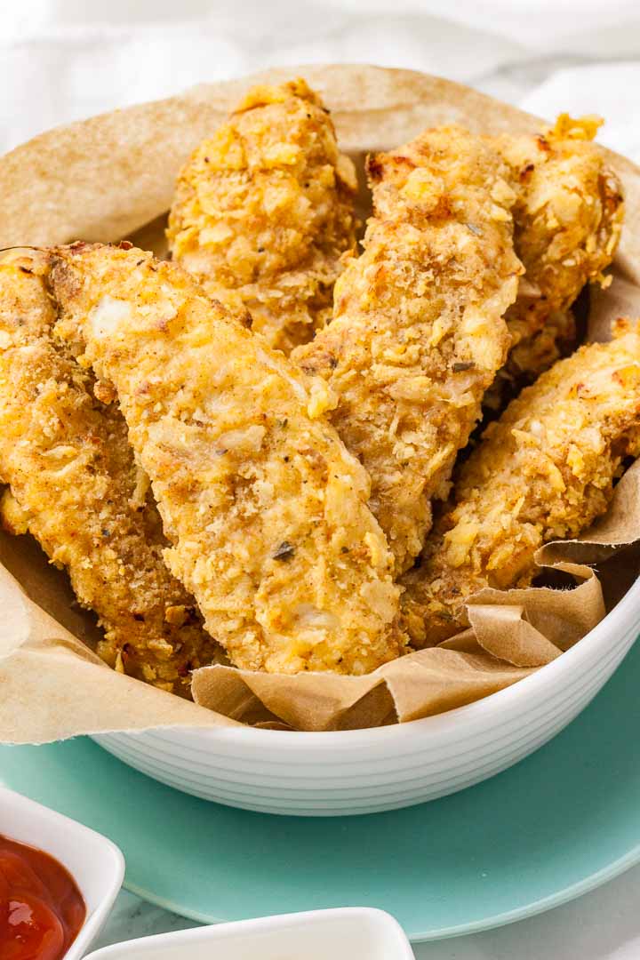 Close-up of a white bowl lined with parchment paper, containing fried chicken tenders stacked on a teal plate.