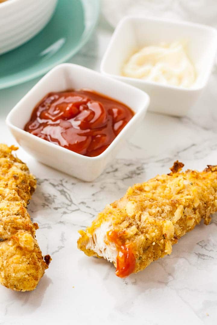 A chicken Tender, with a bite taken out, with some ketchup on a marble surface, next to a whole tender, with small white square bowls of ketchup and mayonaise.