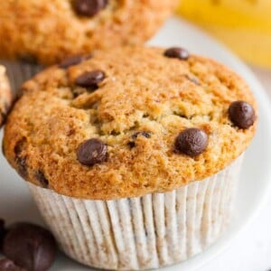 A white plate of banana chocolate chip muffins in front of a banana.
