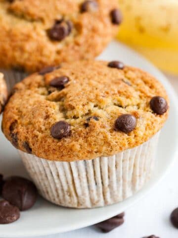 A white plate of banana chocolate chip muffins in front of a banana.